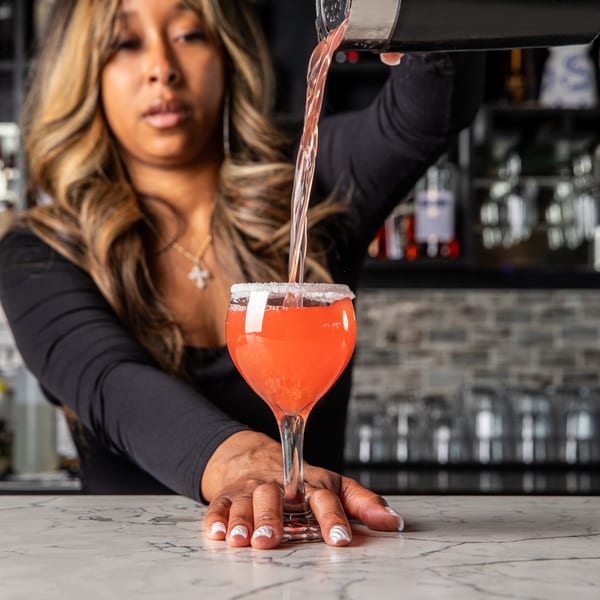A bartender pours a red cocktail from a shaker into a sugar-rimmed glass on a marble bar.