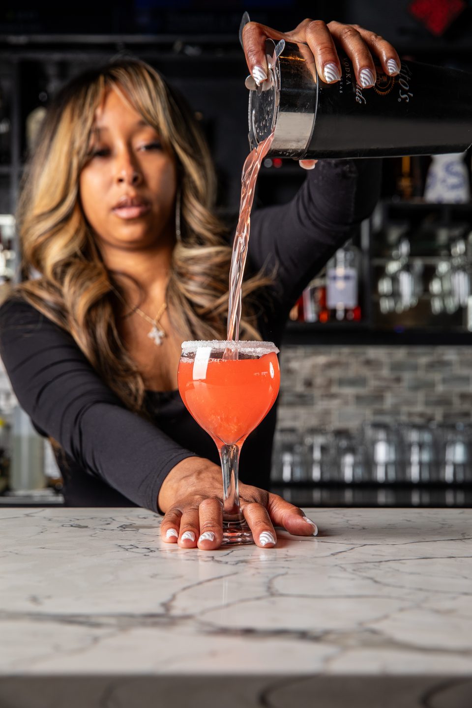 A bartender pours a red cocktail from a shaker into a sugar-rimmed glass on a marble bar.