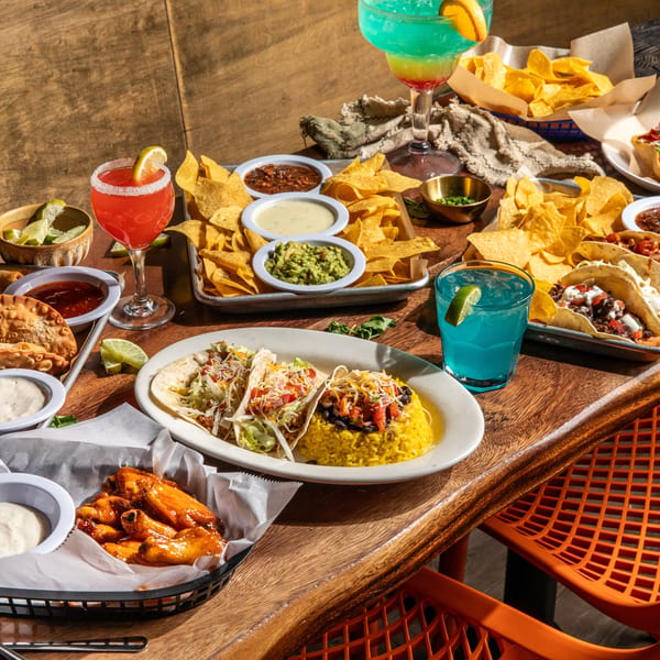 A wooden table laden with various Mexican dishes, including tacos, nachos, empanadas, and colorful drinks.