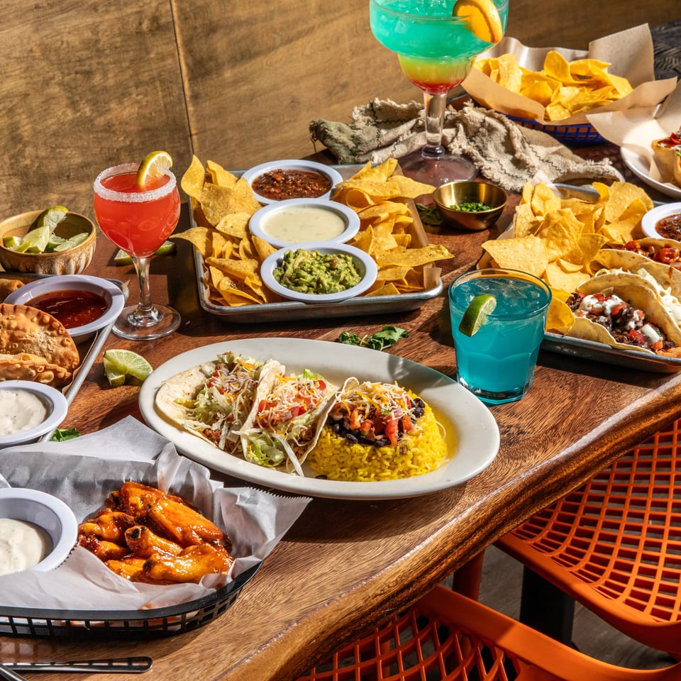 A wooden table laden with various Mexican dishes, including tacos, nachos, empanadas, and colorful drinks.