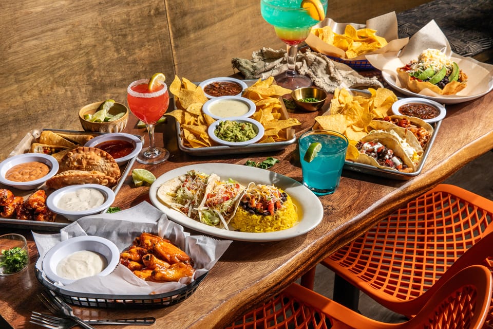 A wooden table laden with various Mexican dishes, including tacos, nachos, empanadas, and colorful drinks.