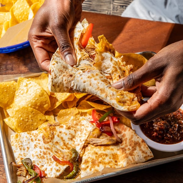 Hands holding a quesadilla slice filled with chicken and peppers, with chips and salsa on a tray.