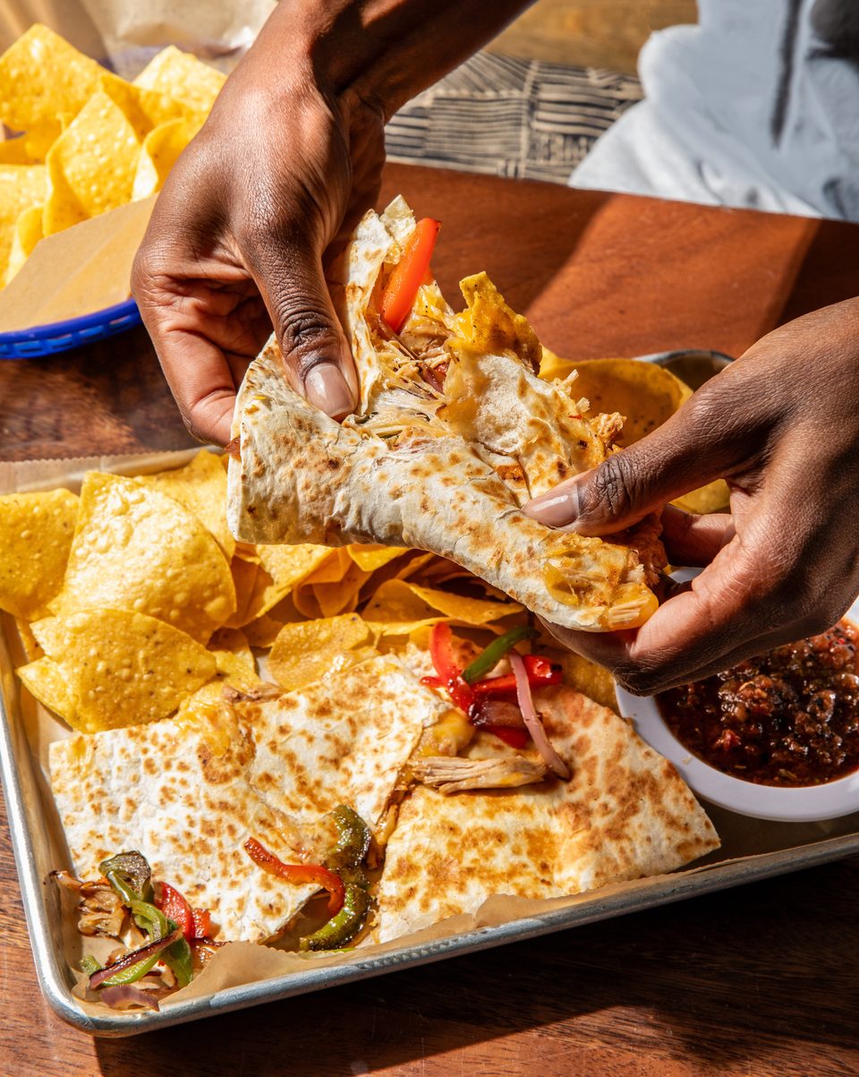 Hands holding a quesadilla slice filled with chicken and peppers, with chips and salsa on a tray.