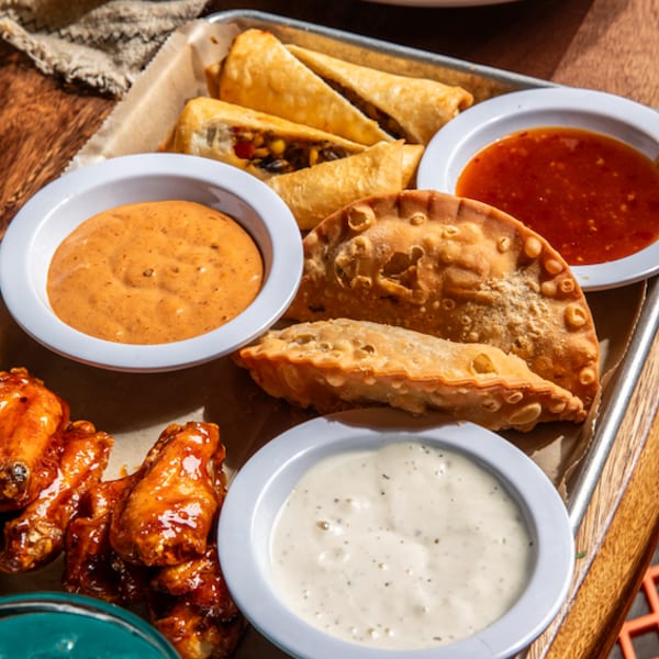 A tray of appetizers including empanadas, taquitos, chicken wings, and three dipping sauces.