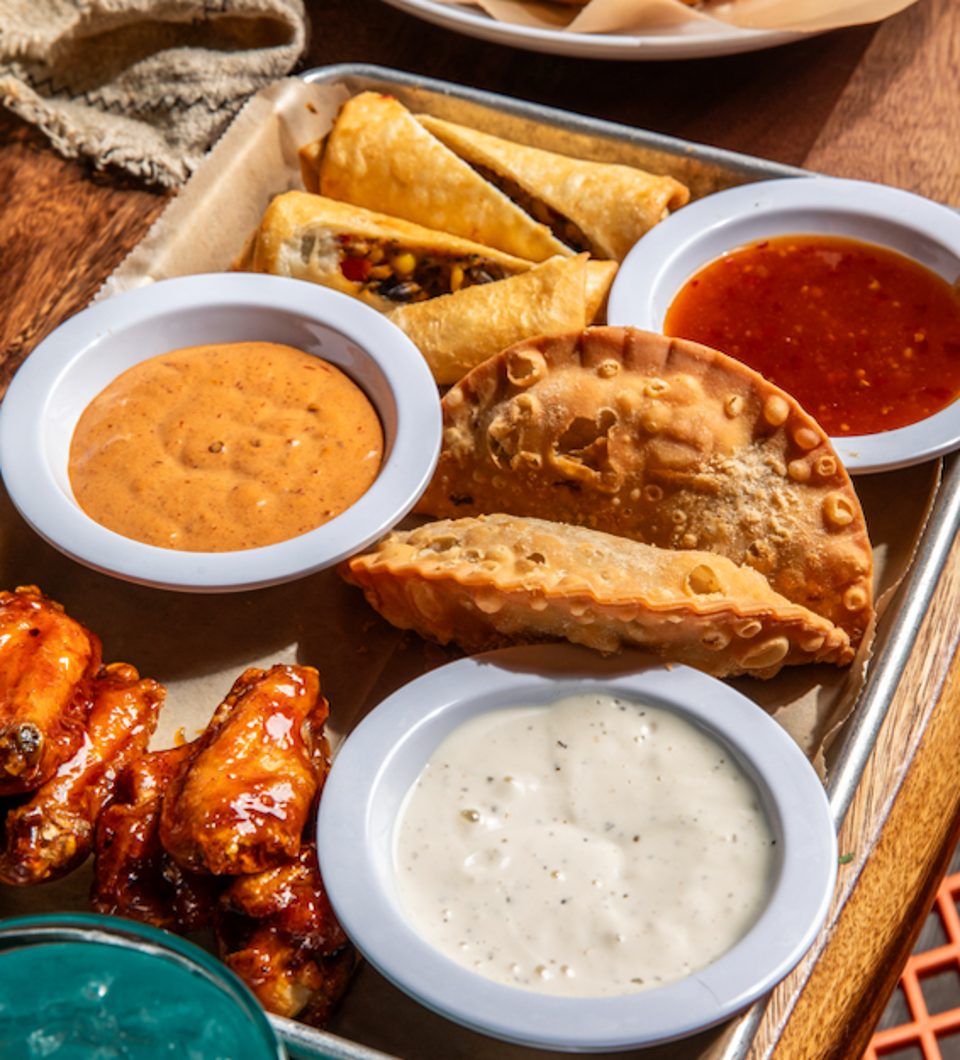 A tray of appetizers including empanadas, taquitos, chicken wings, and three dipping sauces.