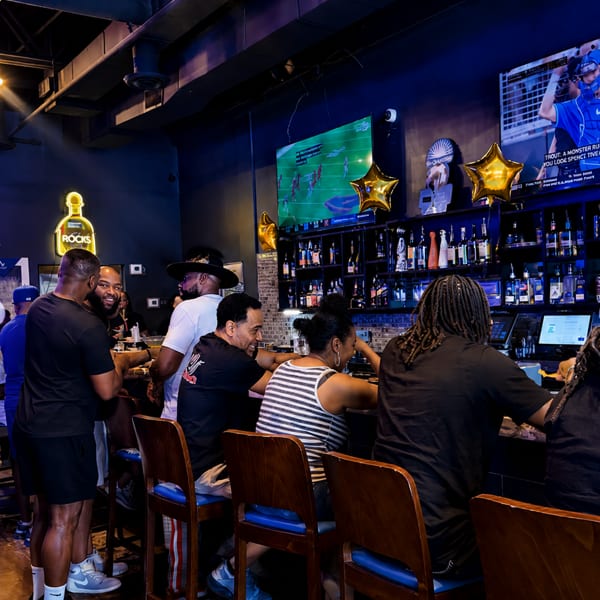 Patrons socialize at a dark, lively bar with glowing screens and a neon "Rocks" sign.