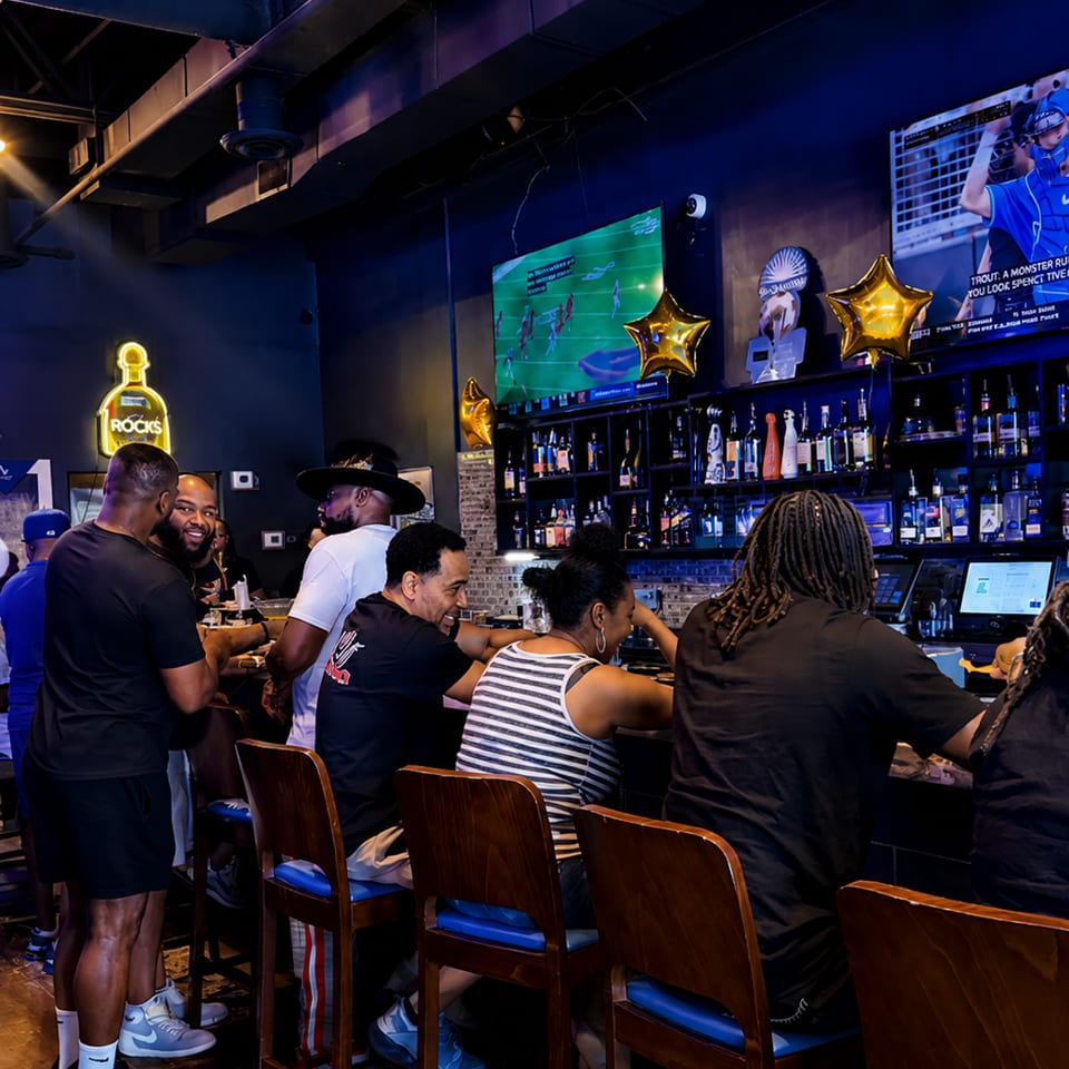 Patrons socialize at a dark, lively bar with glowing screens and a neon "Rocks" sign.
