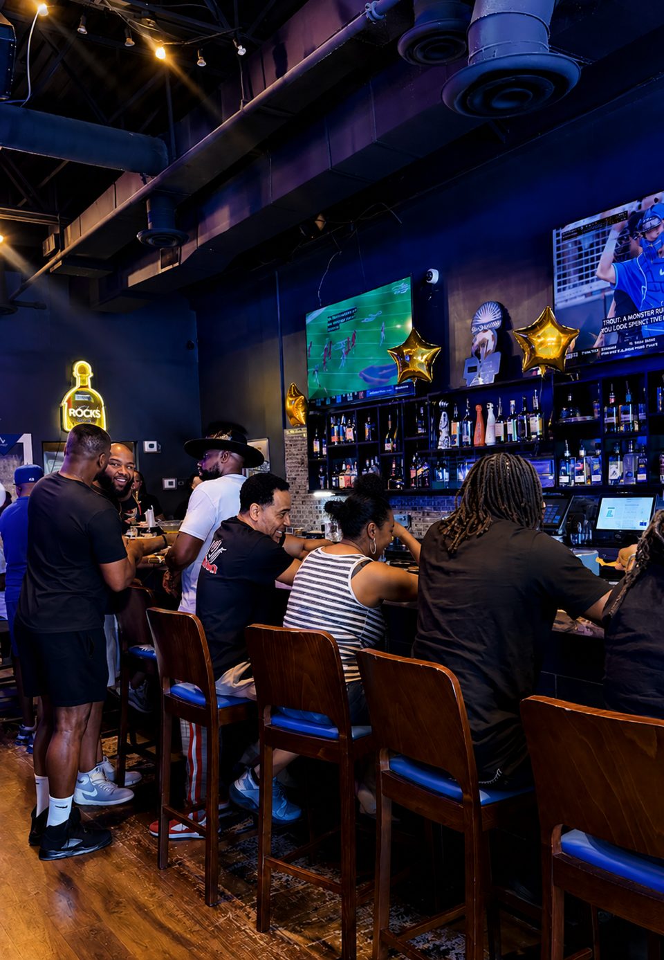 Patrons socialize at a dark, lively bar with glowing screens and a neon "Rocks" sign.