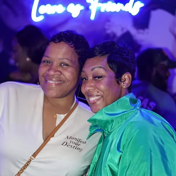 Two smiling women pose for a photo under a neon sign that reads "Love or Friends".