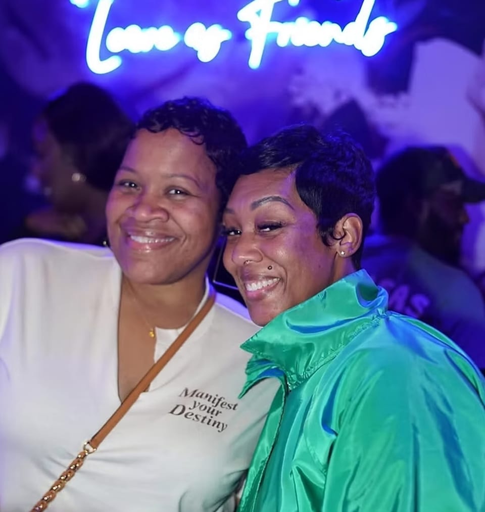 Two smiling women pose for a photo under a neon sign that reads "Love or Friends".