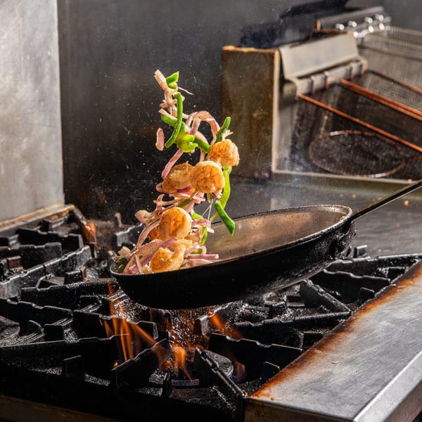Shrimp, green peppers, and red onions are tossed in a pan over a fiery stove.
