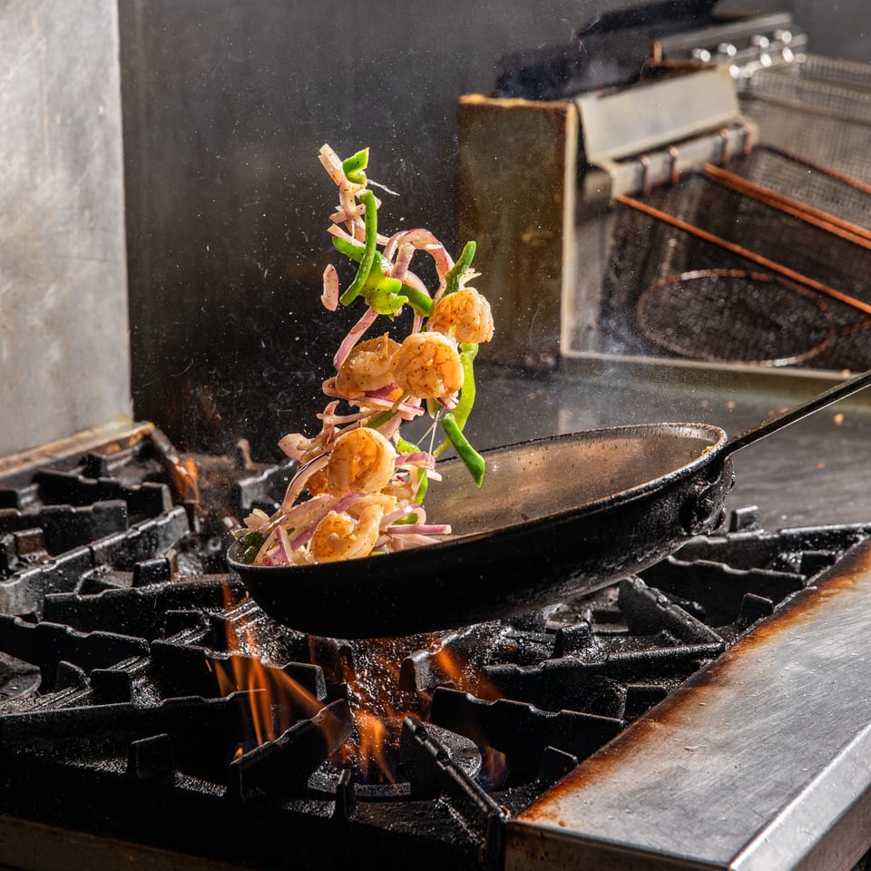 Shrimp, green peppers, and red onions are tossed in a pan over a fiery stove.