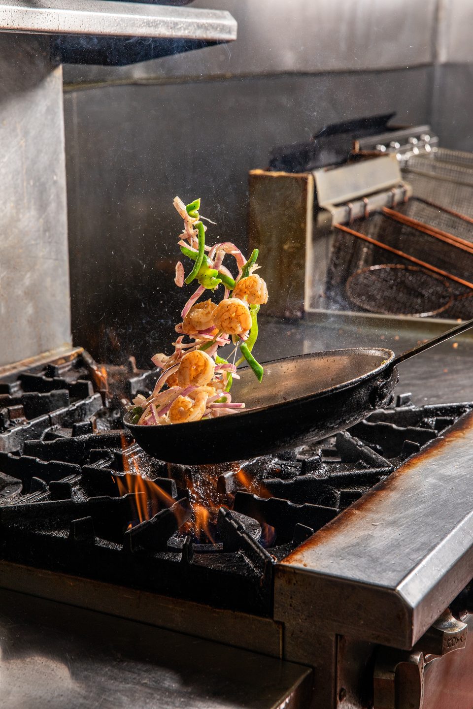 Shrimp, green peppers, and red onions are tossed in a pan over a fiery stove.