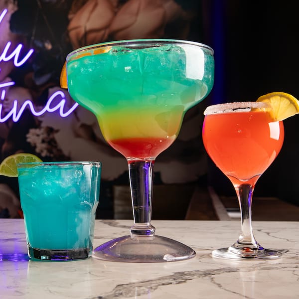 Three colorful cocktails, including a large layered drink, on a marble bar with a neon sign.
