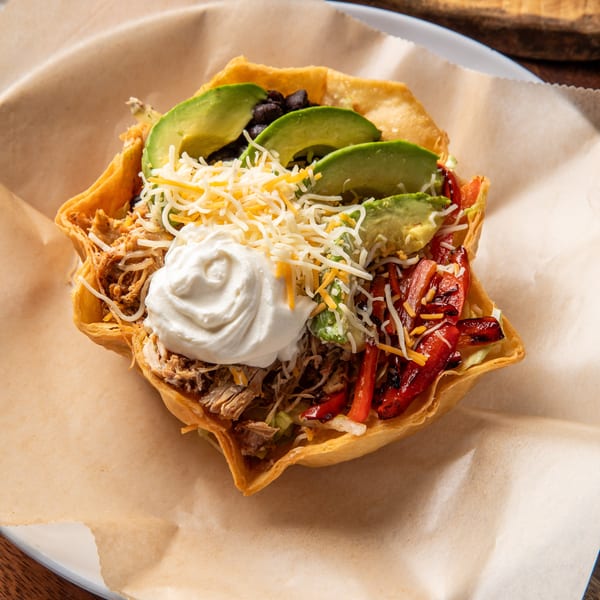 A taco salad in a crispy tortilla bowl with pulled pork, avocado, cheese, and sour cream.