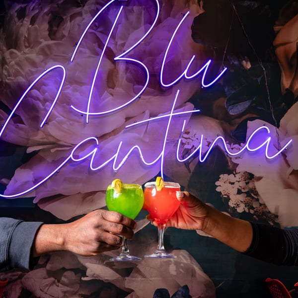 Two hands clinking green and red margaritas in front of a purple neon "Blu Cantina" sign.