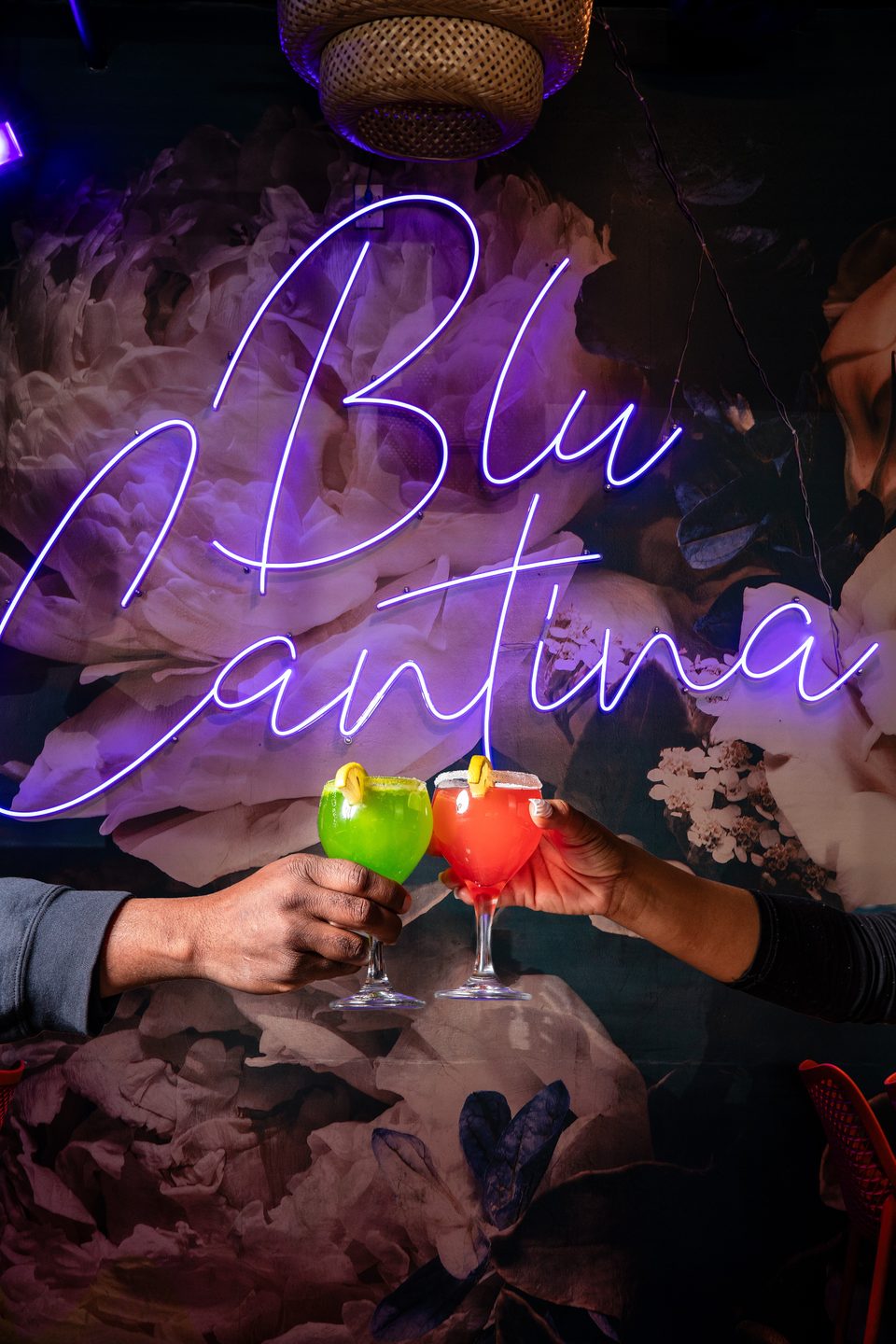 Two hands clinking green and red margaritas in front of a purple neon "Blu Cantina" sign.