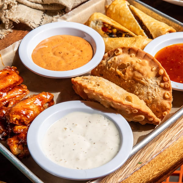 A tray of appetizers including glazed chicken wings, empanadas, and spring rolls with dipping sauces.