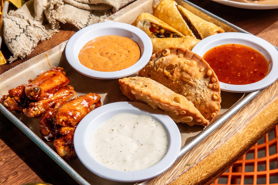 A tray of appetizers including glazed chicken wings, empanadas, and spring rolls with dipping sauces.