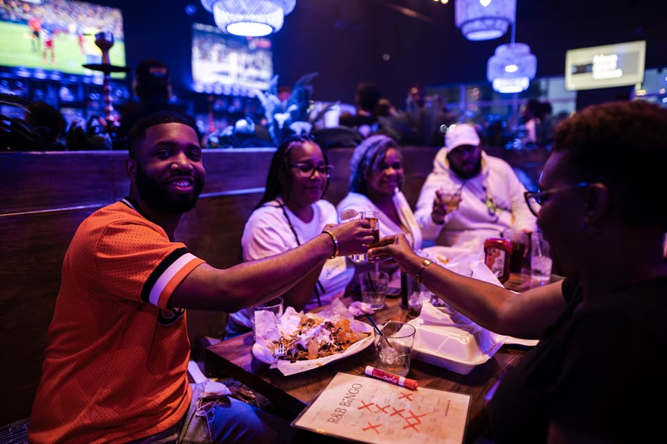 Friends toast drinks at a table with nachos and an R&B Bingo card in a dimly lit restaurant.