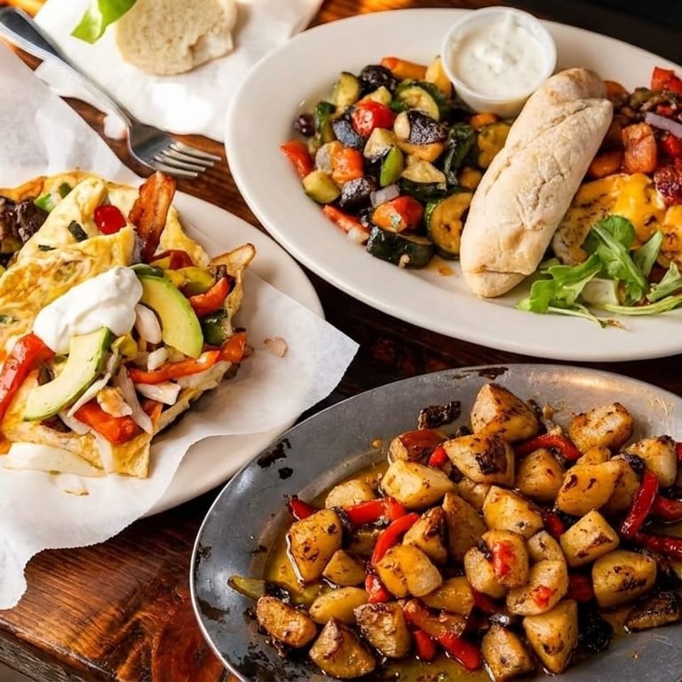 Three plates of breakfast food, including omelets, roasted vegetables, and potatoes, on a wooden table.