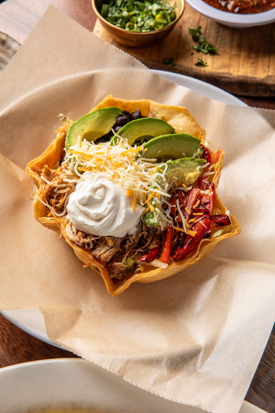Taco salad in a crispy tortilla bowl, topped with shredded chicken, avocado, sour cream, and cheese.