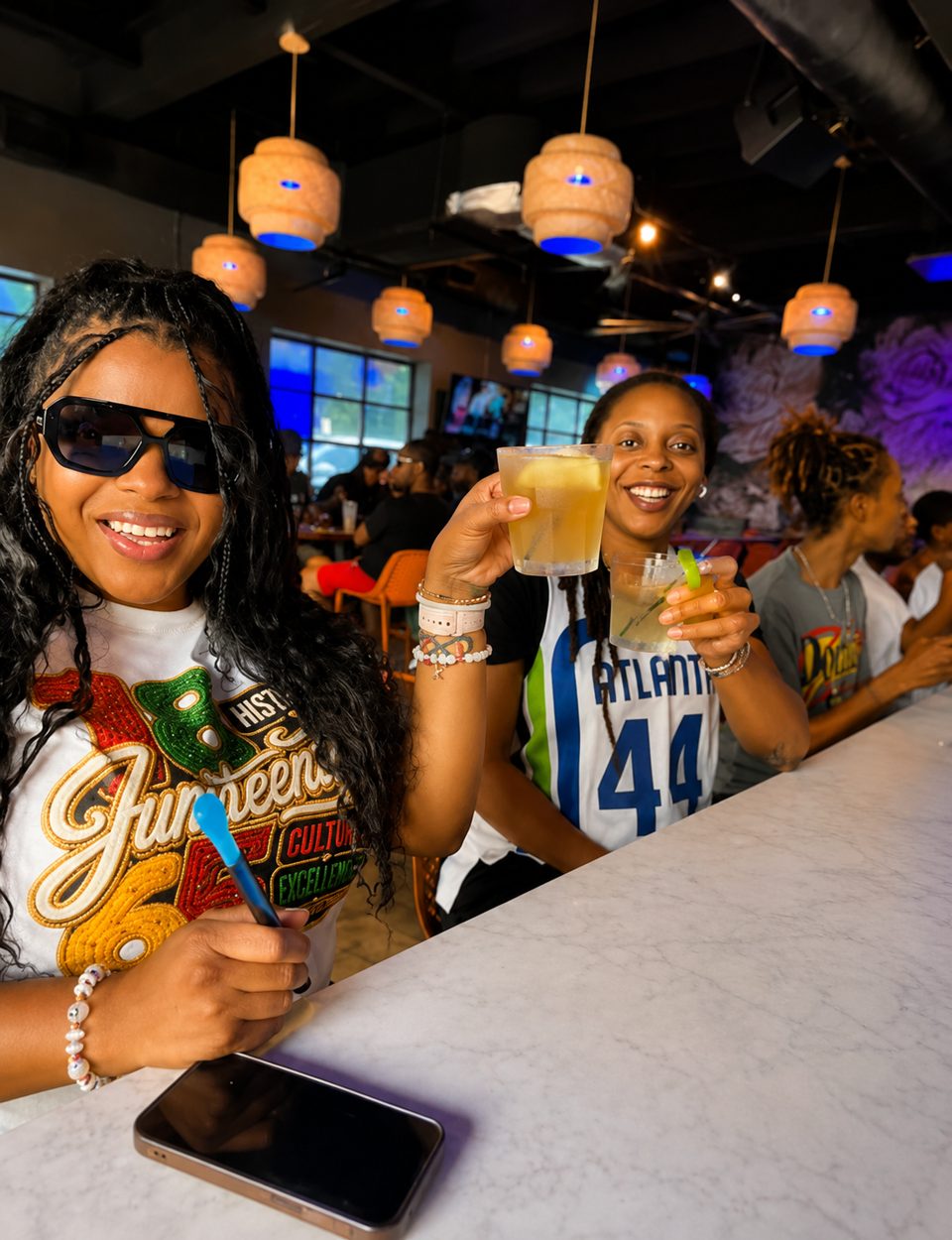 Two women smiling and toasting drinks at a lively bar with unique lighting and murals.
