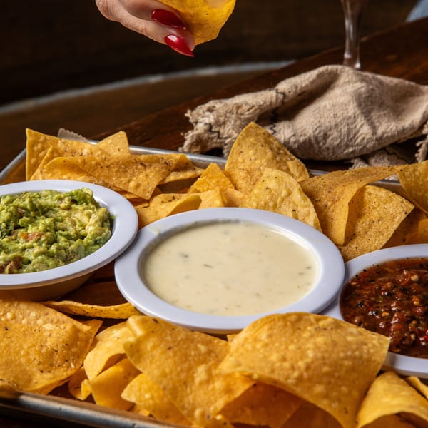 A hand with red nails dips a tortilla chip into a bowl of queso, with guacamole and salsa nearby.
