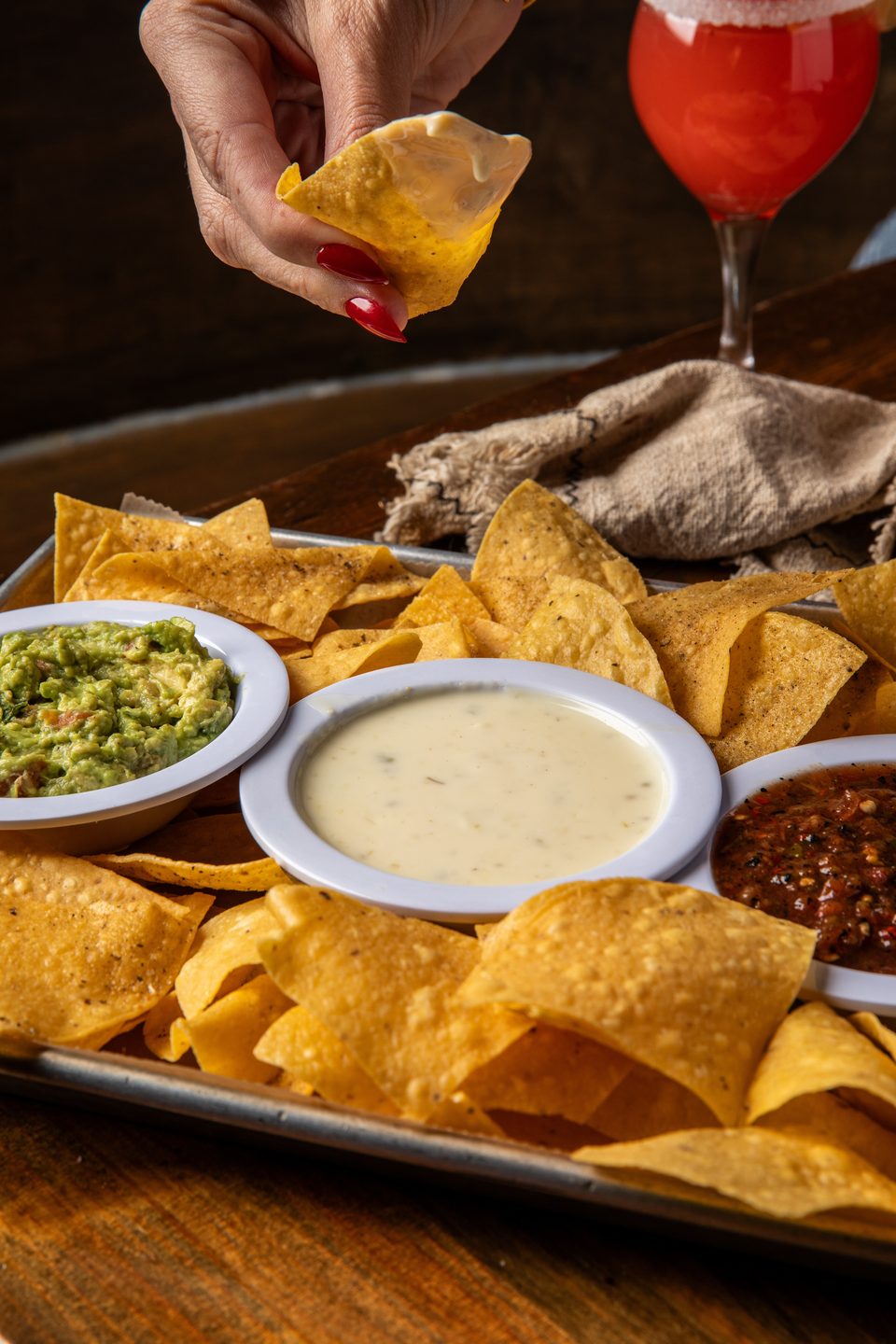 A hand with red nails dips a tortilla chip into a bowl of queso, with guacamole and salsa nearby.
