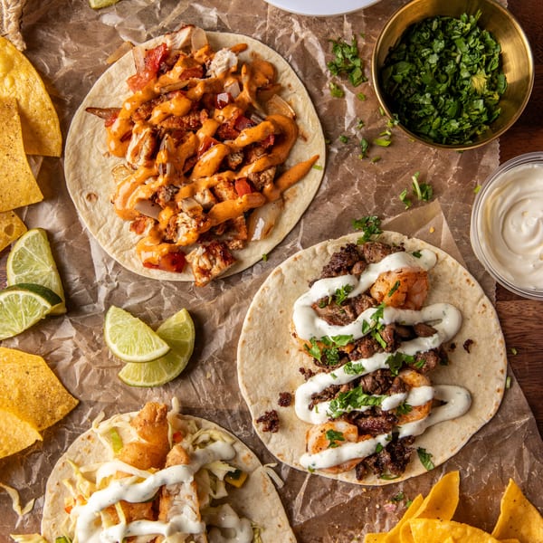 Three vibrant tacos, salsa, cilantro, and tortilla chips arranged on crumpled brown paper.