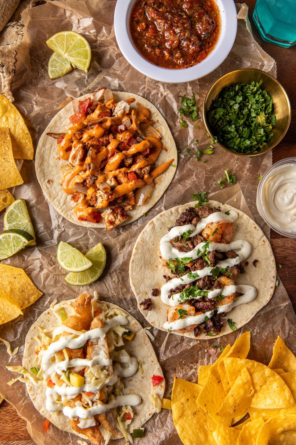 Three vibrant tacos, salsa, cilantro, and tortilla chips arranged on crumpled brown paper.