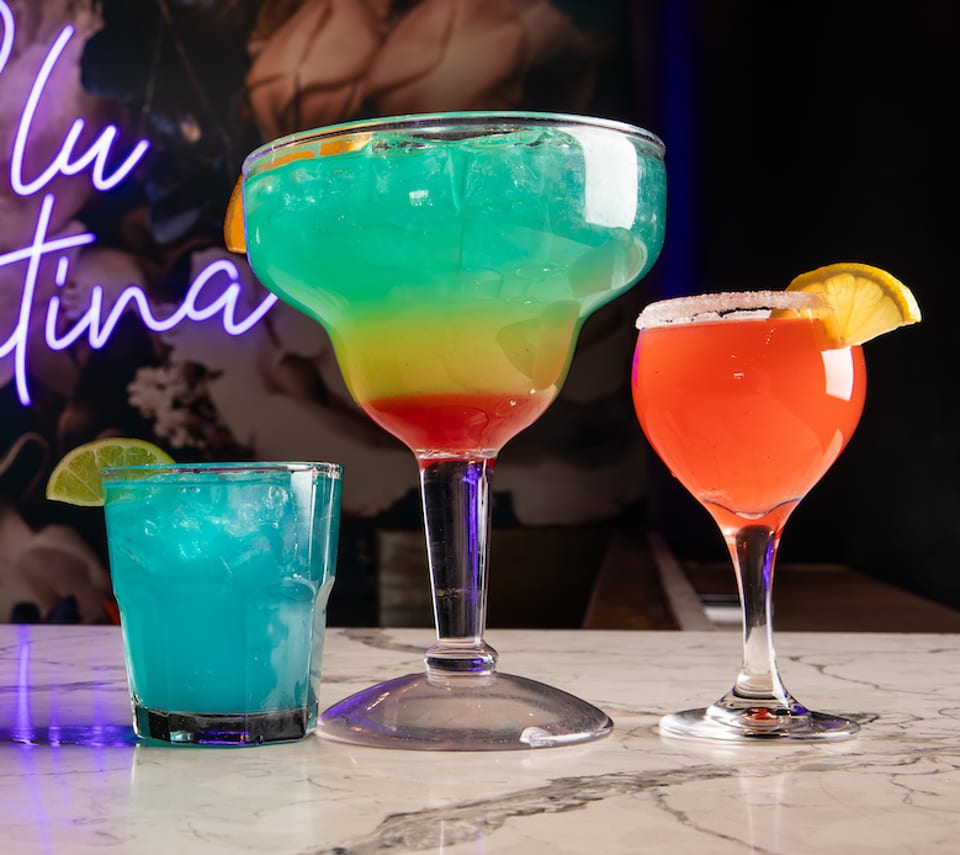 Three colorful cocktails, including a large layered drink, on a marble bar with a neon sign.