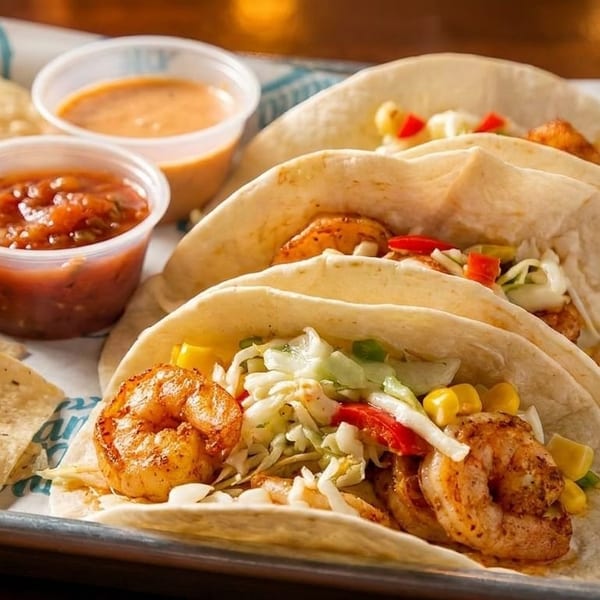 Three shrimp tacos with cabbage, corn, and peppers, served with chips, salsa, and sauce.