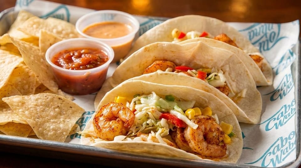 Three shrimp tacos with cabbage, corn, and peppers, served with chips, salsa, and sauce.