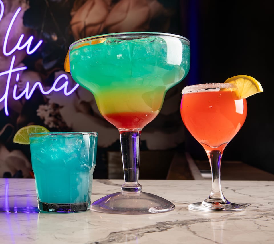 Three colorful cocktails, including a large layered drink, on a marble bar with a neon sign.