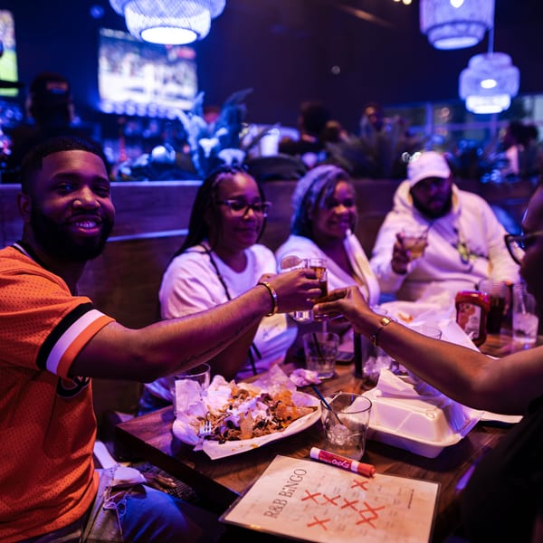 Friends clinking glasses at a table with nachos and an R&B Bingo card in a dimly lit restaurant.