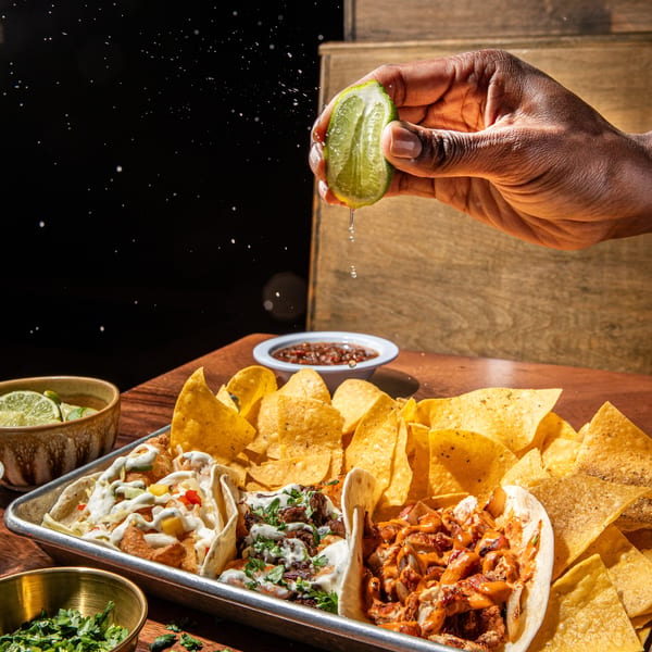 A hand squeezes a lime over a tray of tacos, tortilla chips, and salsa on a wooden table.
