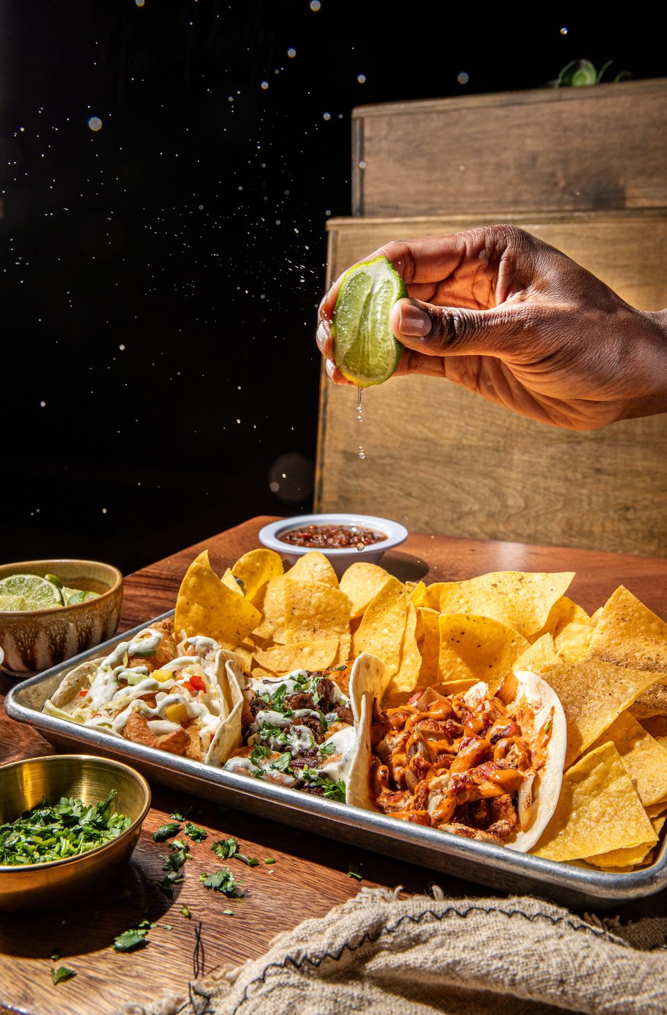 A hand squeezes a lime over a tray of tacos, tortilla chips, and salsa on a wooden table.