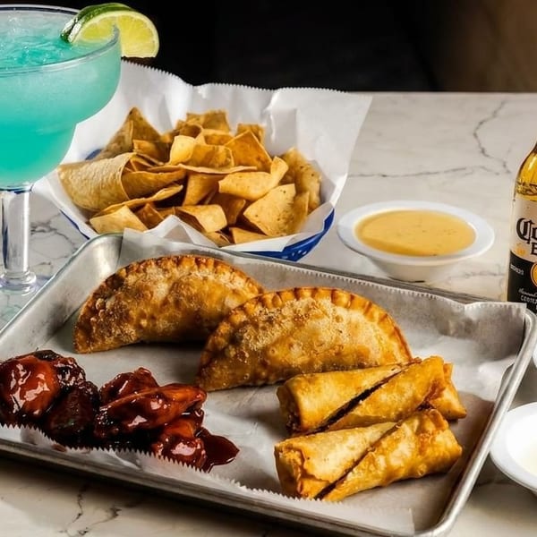A blue margarita, Corona beer, chips, empanadas, wings, and egg rolls with dipping sauces.