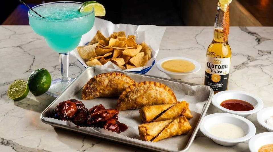 A blue margarita, Corona beer, chips, empanadas, wings, and egg rolls with dipping sauces.